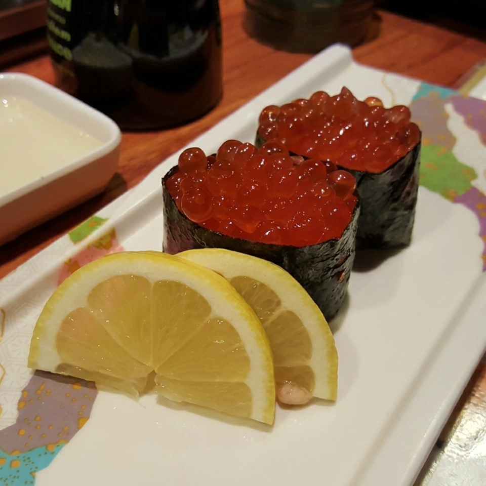 Two ikura sushi pieces topped with salmon roe sit on a decorative plate beside lemon slices, dipping sauce, and a sushi menu.